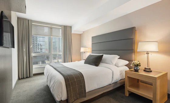 Quarto claro com cama branca e janela voltada para os prédios da cidade